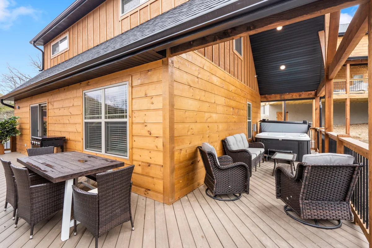 A wooden deck with an inviting outdoor seating area and a private hot tub, set against a scenic backdrop of the Ozark hills at sunset. The space is arranged for al fresco dining and relaxation, with lush surroundings enhancing the tranquil, romantic atmosphere.