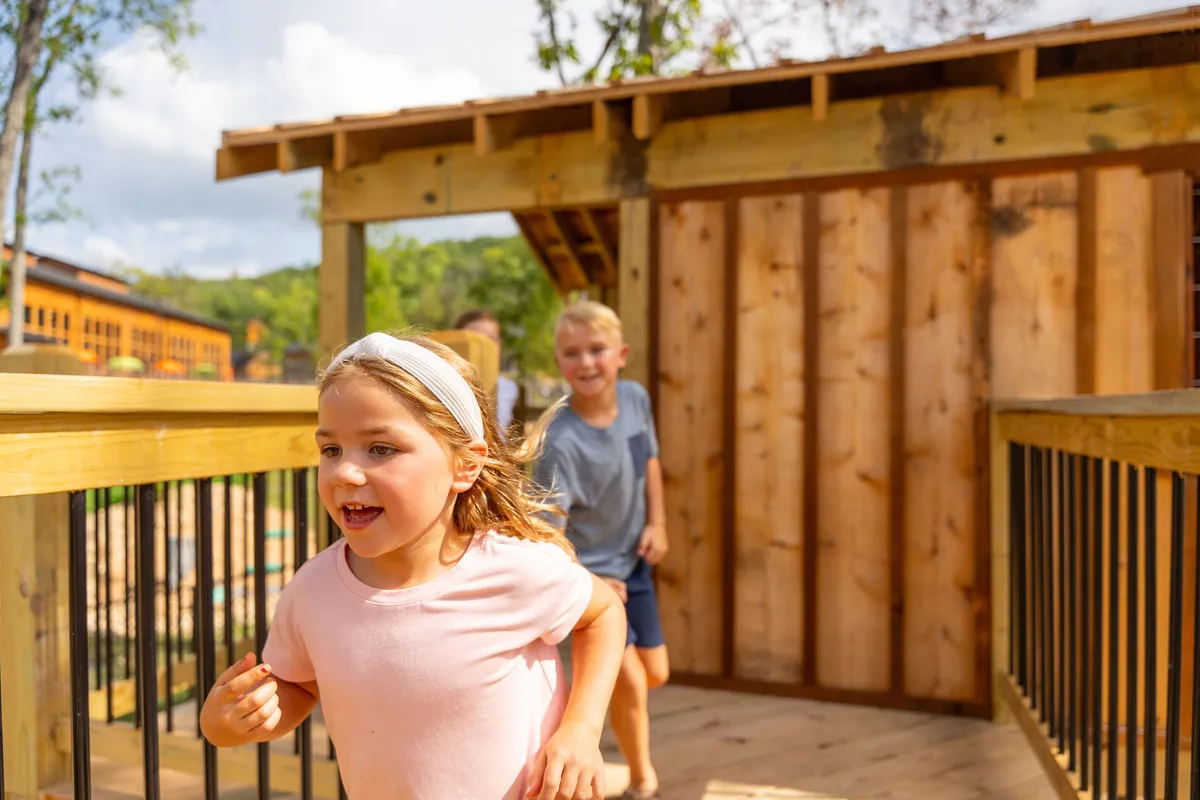 Children joyfully play on a wooden structure surrounded by lush greenery. The scene is filled with energy and laughter, capturing the spirit of outdoor exploration and imaginative play.