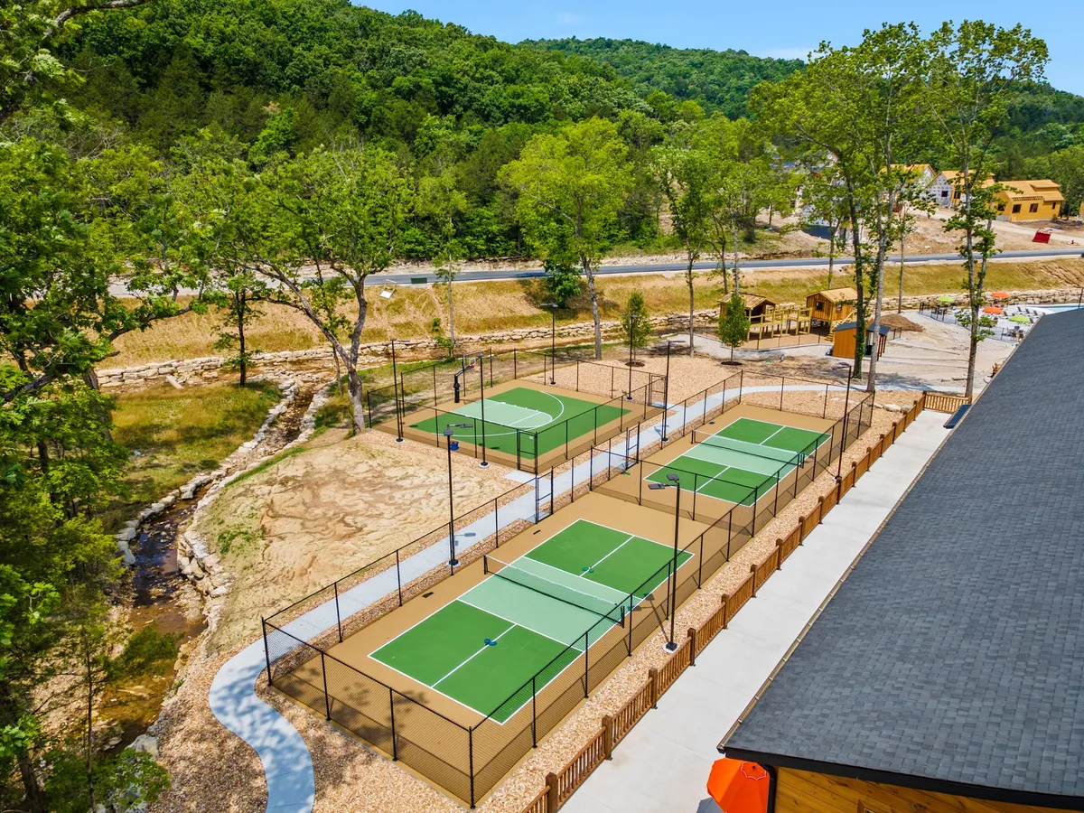 An exterior view of well-maintained tennis courts with a walking path winding beside them, surrounded by tall trees and a nearby playground area. The scene is active yet relaxed, with plenty of space for family activities and friendly games.