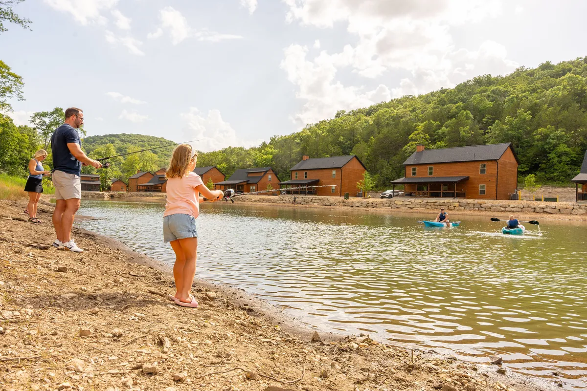 Families are gathered by a lake, with children fishing by the water, adults casting lines, and kayakers paddling nearby. Wooden cabins are visible in the background, set amidst lush greenery, creating a sense of adventure and community.