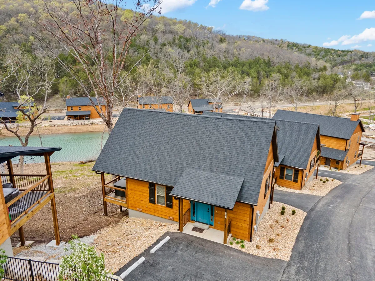 A cluster of wooden cabins with vibrant blue doors nestled beside clear water, surrounded by lush greenery and inviting outdoor spaces. The setting hints at nearby hiking trails and areas for social gatherings or barbecues.