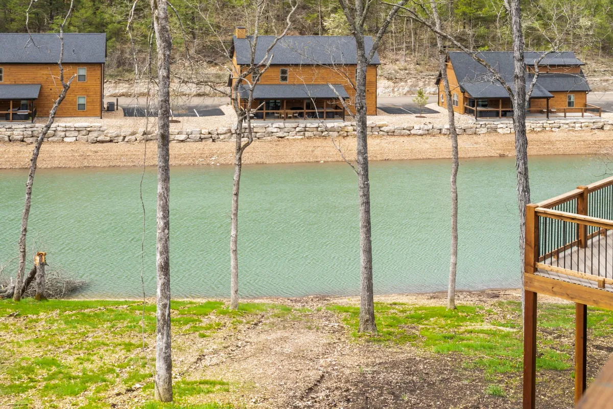 A tranquil riverside scene featuring rustic wooden cabins nestled among trees, with the calm river reflecting the greenery. The setting feels secluded and serene, ideal for relaxation or fishing, and offers a strong sense of connection to the outdoors.