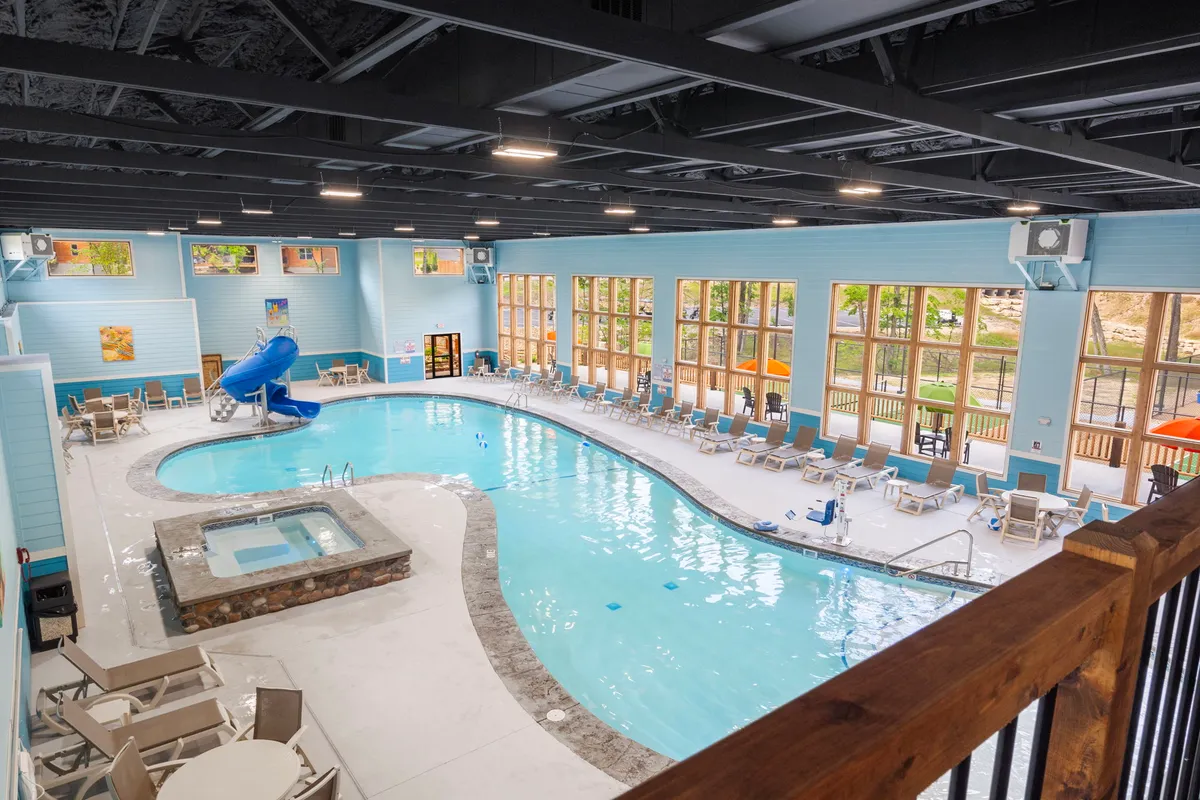 This image features a spacious indoor swimming pool area, complete with a water slide, hot tub, large windows that let in natural light, and several lounge chairs arranged around the pool. People are seen swimming, relaxing, and enjoying the amenities.