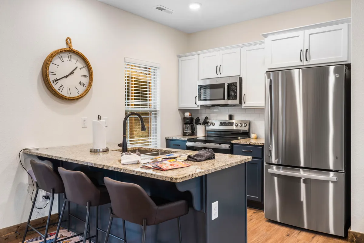 A modern kitchen scene with a sleek granite countertop, contemporary appliances, bar stools for casual seating, and a distinctive clock on the wall. The space is styled for both function and comfort, ideal for cooking or gathering with friends and family.