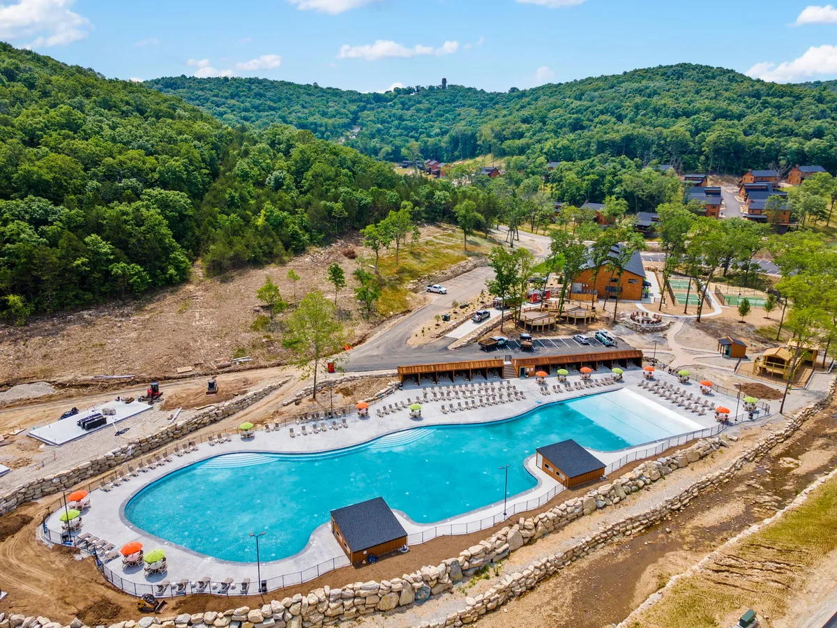 An expansive outdoor swimming pool is surrounded by lush greenery, deck chairs with umbrellas, and nearby recreational structures. The scene is lively and cheerful, suggesting group gatherings and poolside relaxation during warm weather.